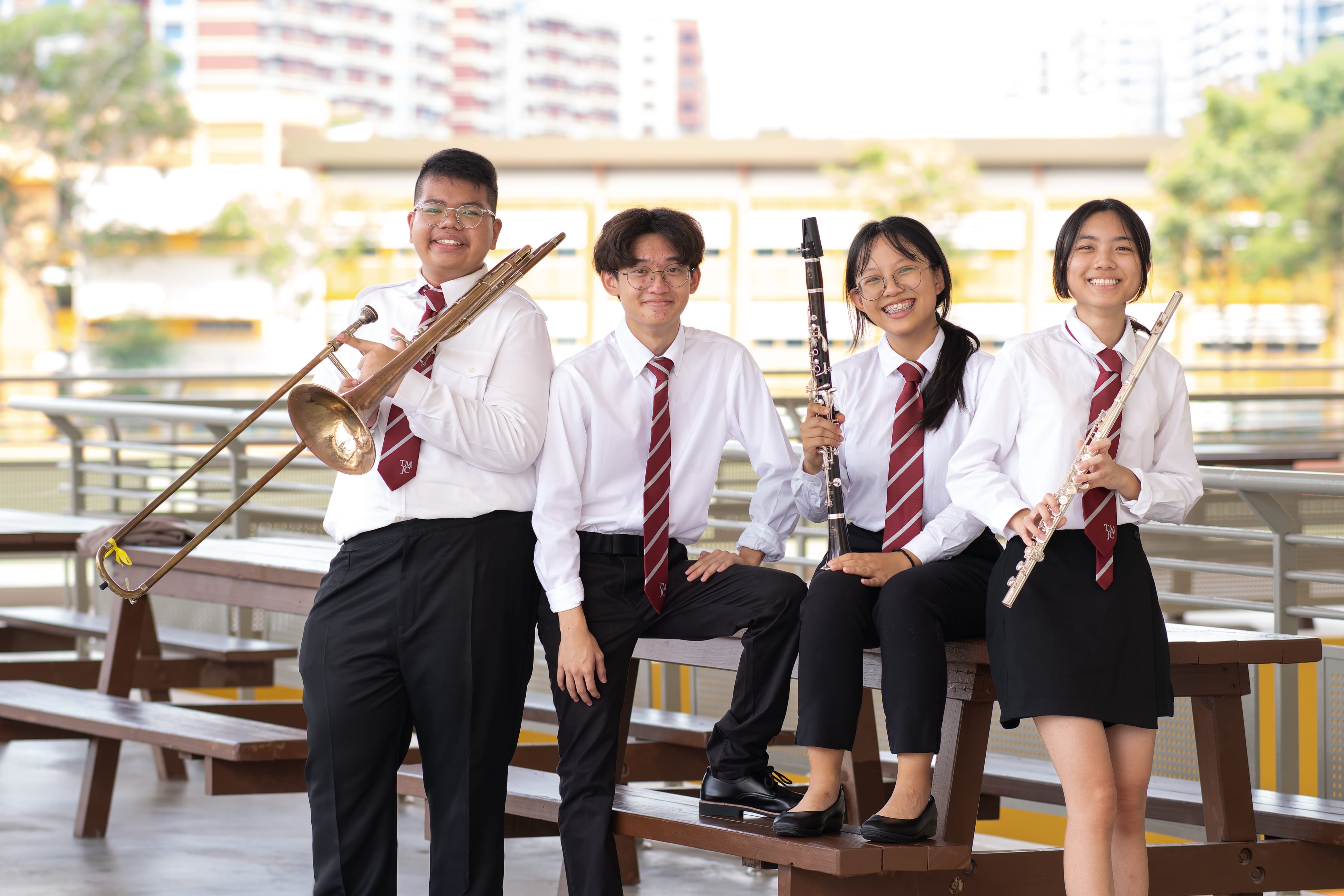 Four smiling students in white school uniforms with red ties pose outdoors, each holding their instrument — a trombone, a clarinet, and a flute — with a urban Singapore backdrop behind them.